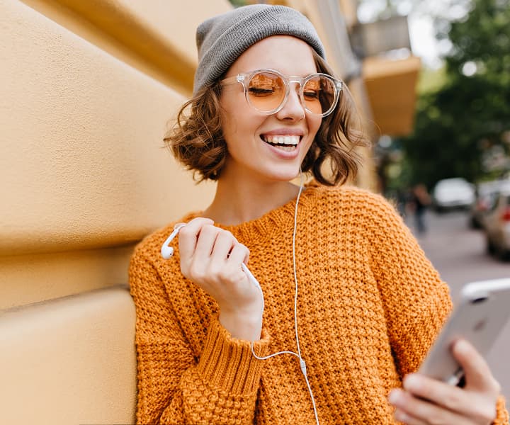 A girl with brown hair, a bob cut, orange glasses, a gray hat, and an orange sweater. One wired earphone in her left ear, the other wired earphone in her right hand, looking at her phone, which she holds in her left hand, and smiling. Standing in a park against the backdrop of an orange fence.